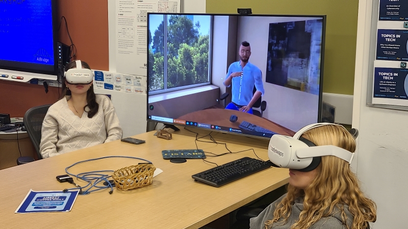 Two female Bentley students sit at a table with a large computer monitor. They are wearing VR headsets and engaging in an immersive learning module from Bodyswaps. 