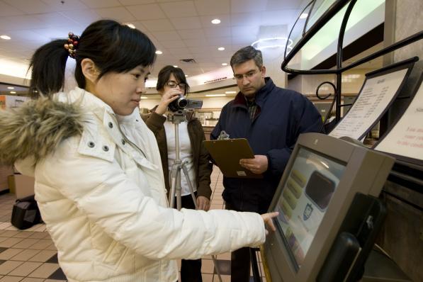 Two researchers observing a woman operating a touch screen kiosk in a shopping center.